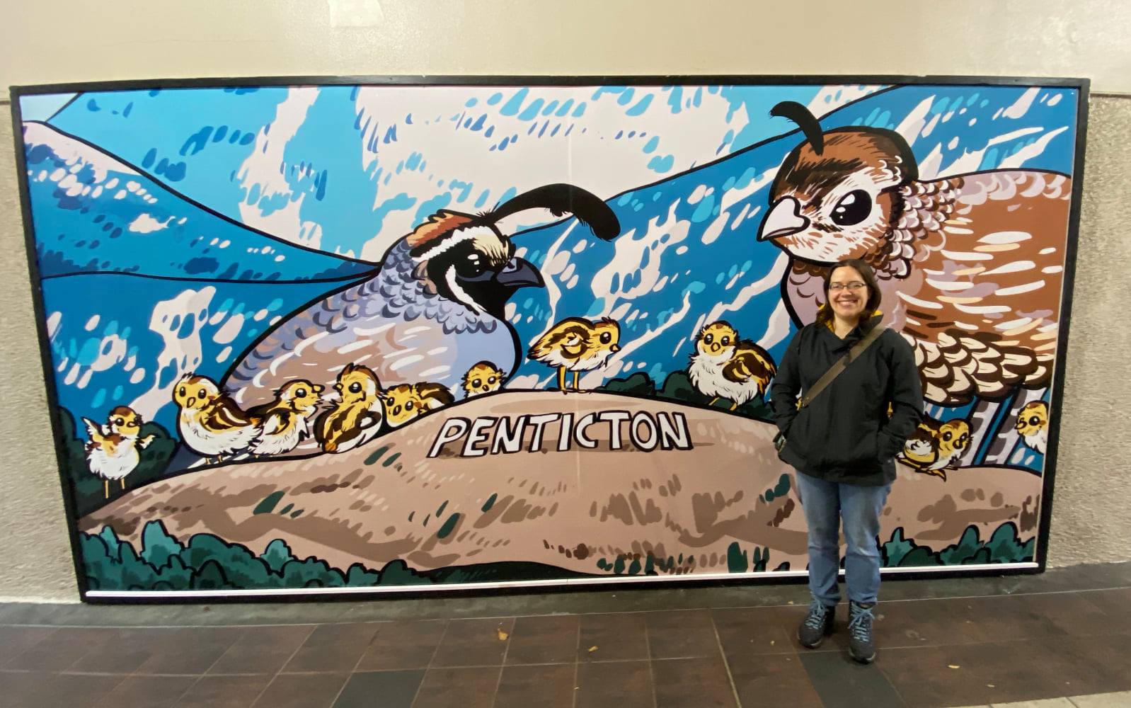 A photo of a woman standing in front of a large mural with large California quails on it.